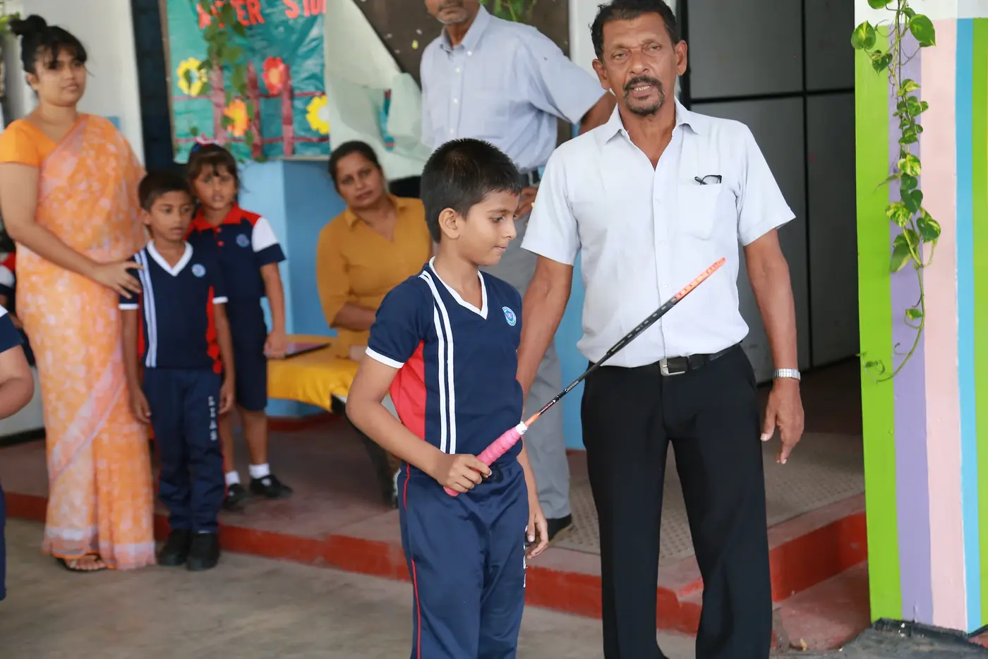 A young boy in sports kit holds a training javelin, with a teacher standing beside him and other children watching.