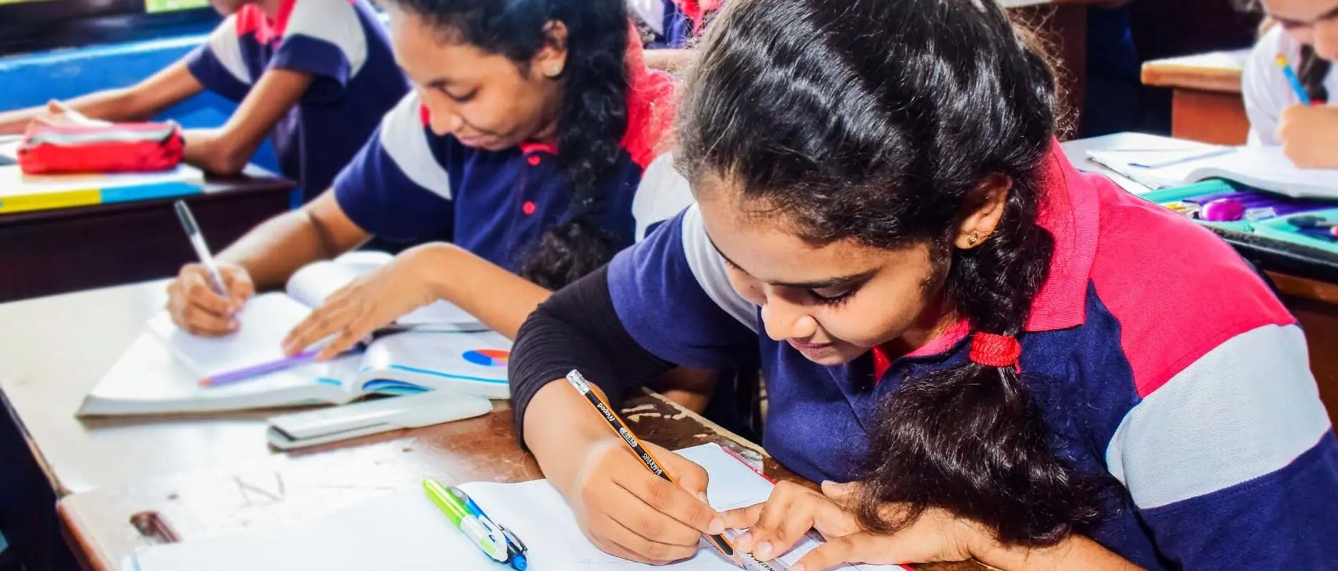 A pupil in the navy, red and white school uniform writes in an exercise book at her desk, with classmates working beside her.