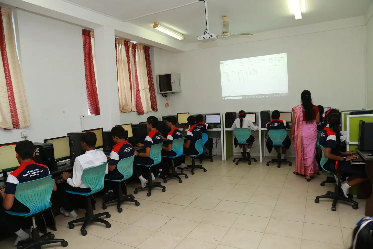 Pupils work at a row of computers while a teacher stands beside a projected spreadsheet.