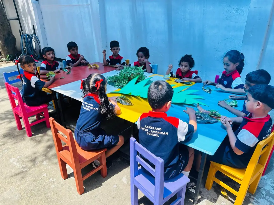 Children sit round a long table outdoors, working with leaves and coloured paper.