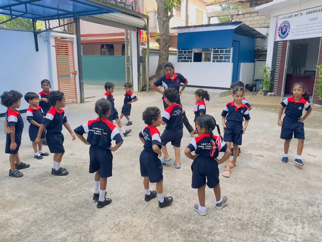 Children stand in the school yard with their hands on their hips, facing a teacher.
