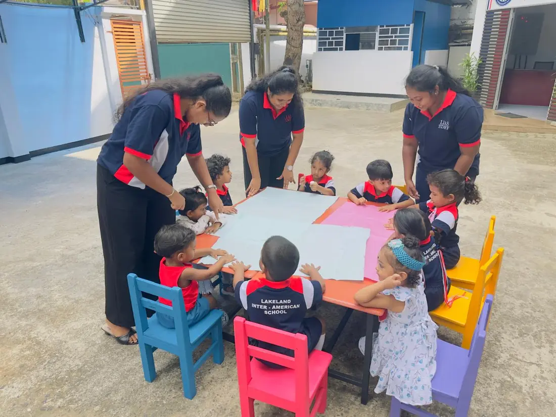 Three teachers help small children spread paper across a table in the yard.