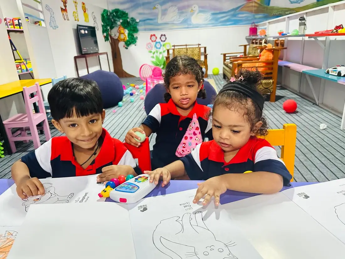 Three small children sit at a table with colouring sheets in a playroom.