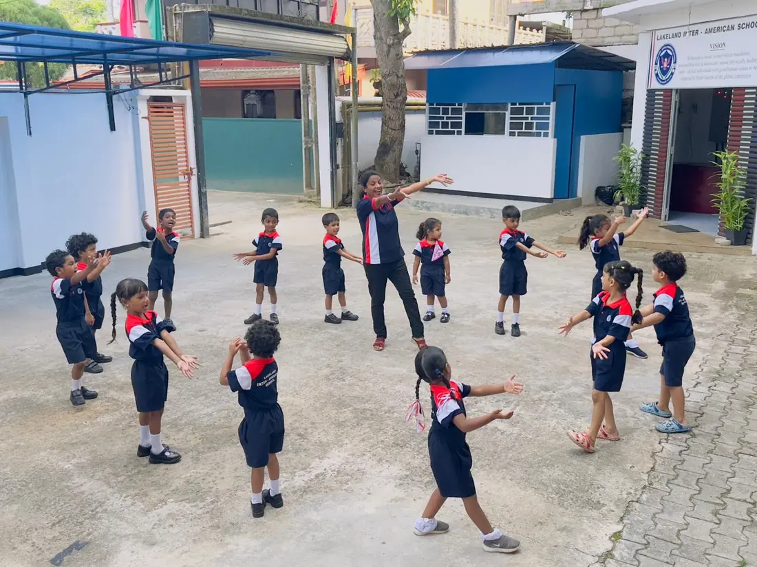 Children stand in a circle in the school yard, copying a teacher who stretches out her arms.