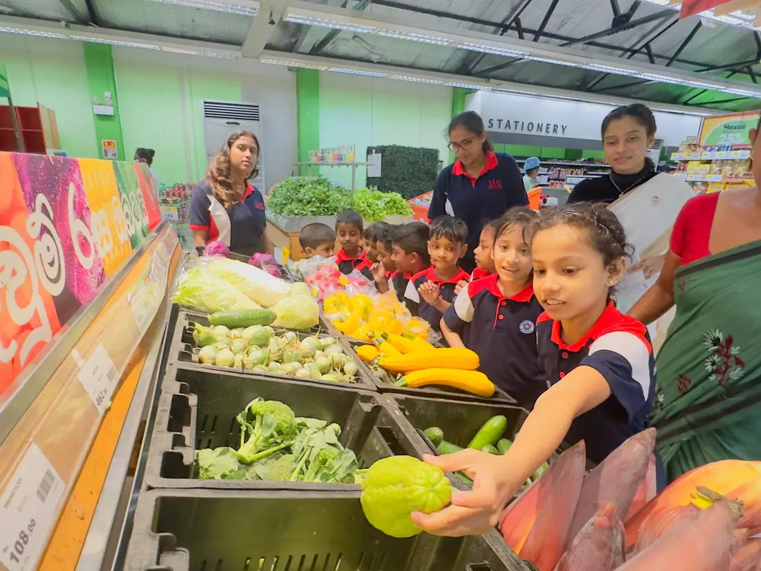 Children on a class visit to a shop look at the vegetables, and one girl reaches for a fruit.