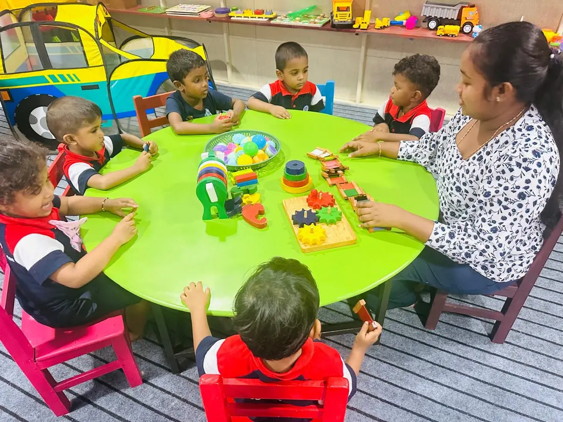 A teacher and small children sit round a green table with wooden puzzles and toys.
