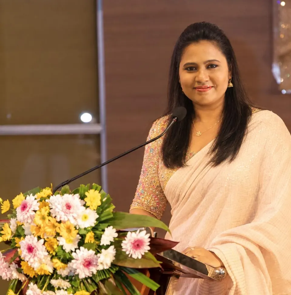 Dr. Nilu Anandappa, in a cream saree, speaking at a lectern decorated with flowers.