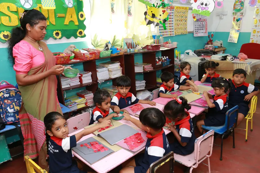A teacher holds a bowl while small children make paper collages at a low table in a classroom.